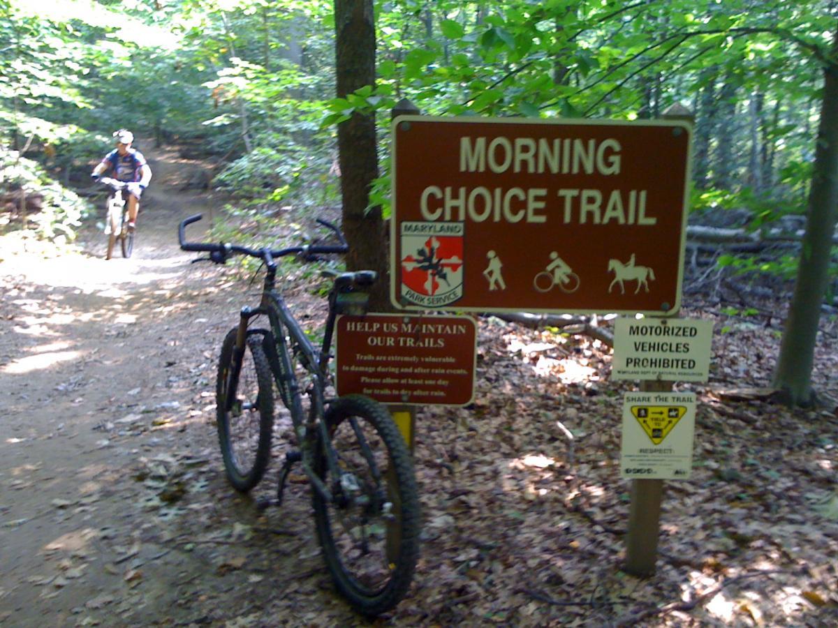 A mountain bike leaning against a trail sign that reads "Morning Choice Trail" in a wooded area, with a cyclist riding on a dirt path in the background. Additional signs nearby emphasize trail maintenance and the prohibition of motorized vehicles. The scene is surrounded by lush green trees and foliage. Patapsco Valley State Park (Avalon Area) mountain bike trail.