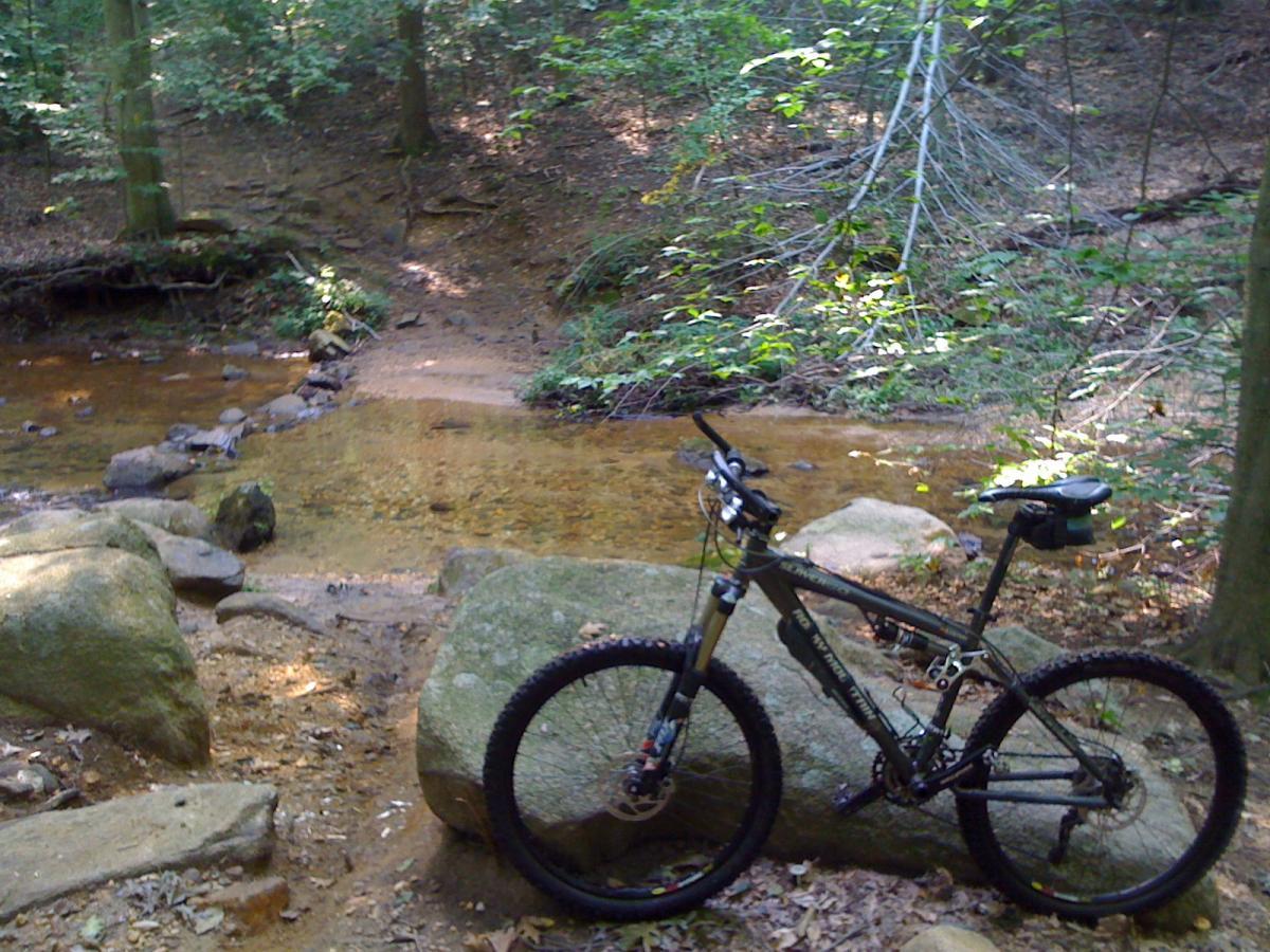 A mountain bike resting on a large rock near a shallow, clear creek surrounded by lush greenery in a wooded area. Sunlight filters through the trees, illuminating the scene. Patapsco Valley State Park (Avalon Area) mountain bike trail.