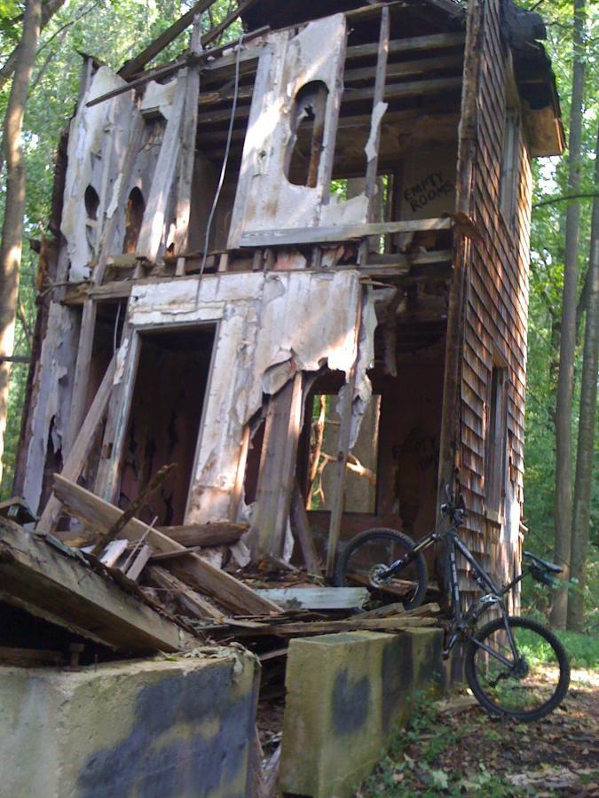 Alt text: An abandoned, partially collapsed wooden house surrounded by trees, with exposed beams and deteriorating walls. A mountain bike is leaning against the structure, and debris is scattered around the base. Patapsco Valley State Park (Avalon Area) mountain bike trail.