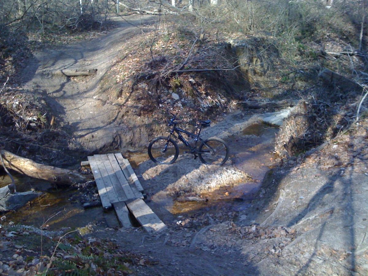 A mountain bike parked on a wooden bridge crossing a small stream in a wooded area. The scene features dirt trails, scattered leaves, and a natural landscape with trees and underbrush. Rosaryville State Park mountain bike trail.