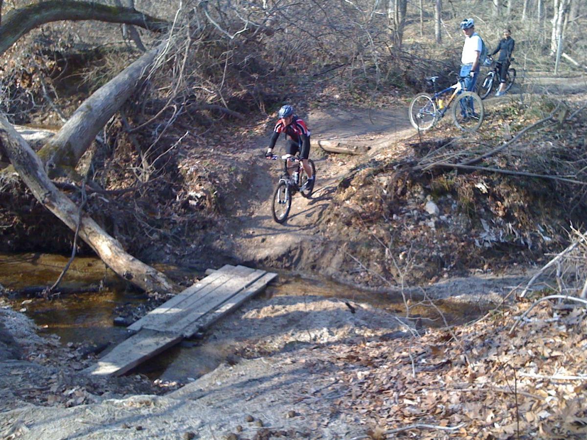 A cyclist rides over a wooden bridge spanning a small creek on a dirt trail surrounded by trees. Two other cyclists are seen in the background, one standing next to their bike while the other is seated on their bicycle, all in a forested area with fallen leaves on the ground. Rosaryville State Park mountain bike trail.