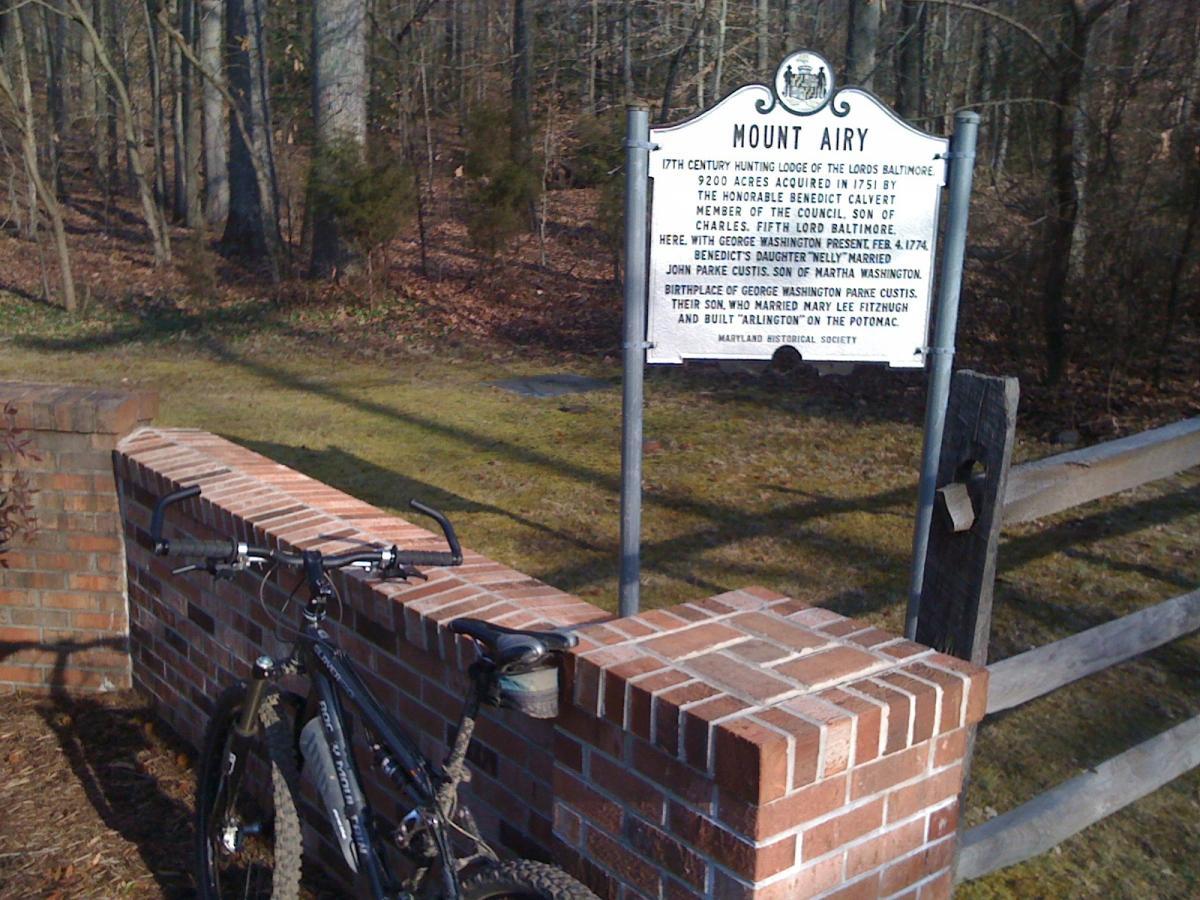 A historical marker for Mount Airy, detailing its significance as a 17th-century hunting lodge of the Lords Baltimore, located in a wooded area. A bicycle is resting against the brick wall of the marker, surrounded by trees and grass. The sign includes information about notable figures such as Benedict Calvert and George Washington. Rosaryville State Park mountain bike trail.
