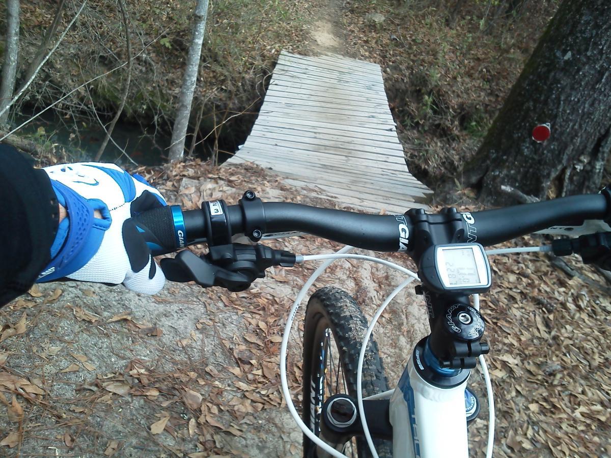 A mountain bike handlebar viewed from the rider's perspective, with one hand gripping the handlebar. In the background, a narrow wooden bridge crosses a small stream, surrounded by autumn leaves and trees. A bike computer on the handlebar displays speed and distance metrics. L.H. Thomson Trails mountain bike trail.