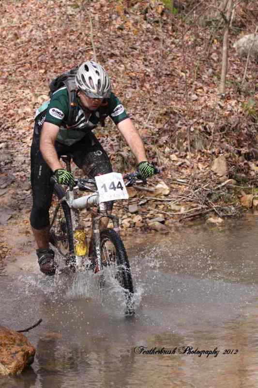 A mountain biker, wearing a helmet and a black and green jersey, is riding through a stream, splashing water as he navigates the rocky terrain. The scene is surrounded by autumn foliage. The biker has a race number, 144, prominently displayed on his front.