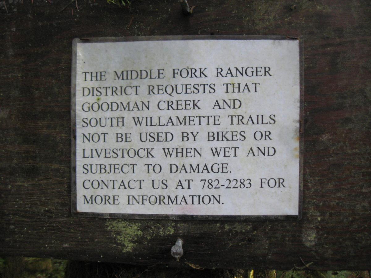 Sign mounted on a wooden post with a notice from the Middle Fork Ranger District. The sign requests that Goodman Creek and South Willamette trails not be used by bikes or livestock when wet, as this may cause damage. It provides a contact number for more information. Hardesty Mountain mountain bike trail.