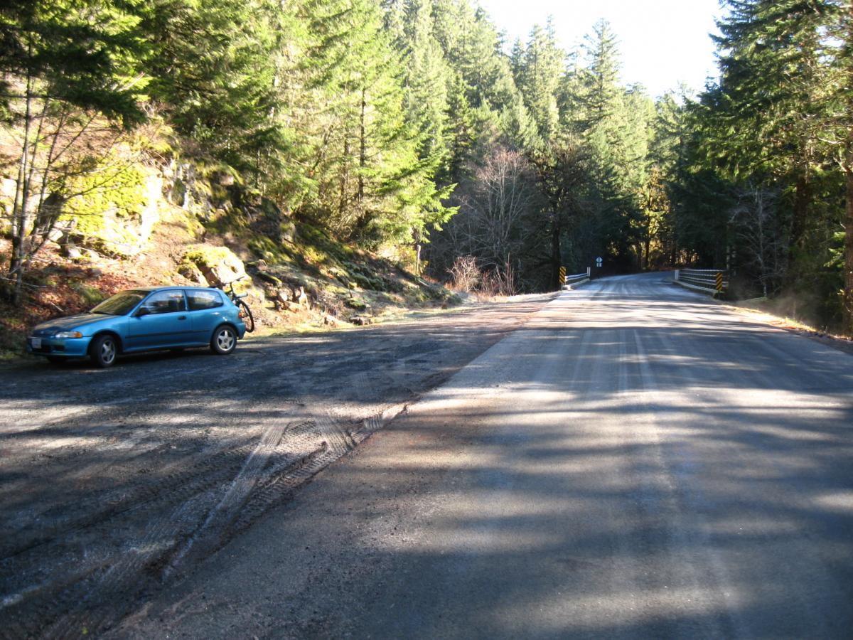 A blue car parked on a gravel shoulder beside a wooded road, surrounded by tall pine trees and rocky terrain. The scene is illuminated by sunlight, casting shadows across the road, which curves gently ahead. Brice Creek mountain bike trail.