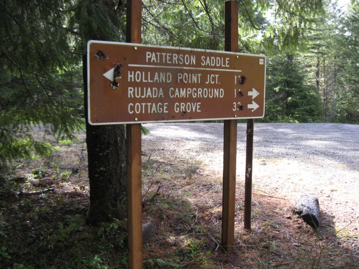 Brown directional sign at a forest intersection indicating distances to Holland Point Junction, Rujada Campground, and Cottage Grove. Surrounded by trees and gravel road. Eula Ridge mountain bike trail.