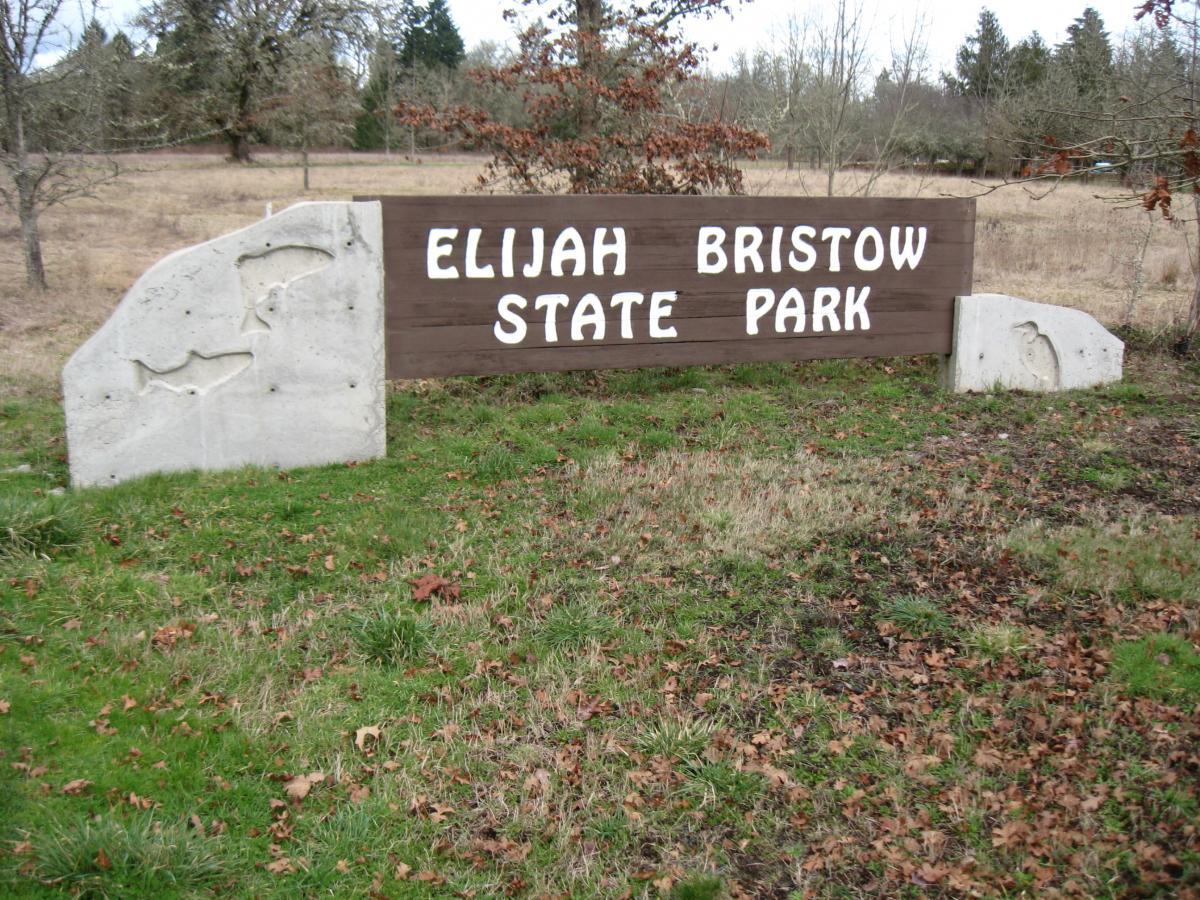 Sign for Elijah Bristow State Park featuring a wooden board with white lettering, surrounded by grass and trees. The sign includes decorative concrete elements carved in the shape of fish and a bird. Elijiah Bristow State Park mountain bike trail.