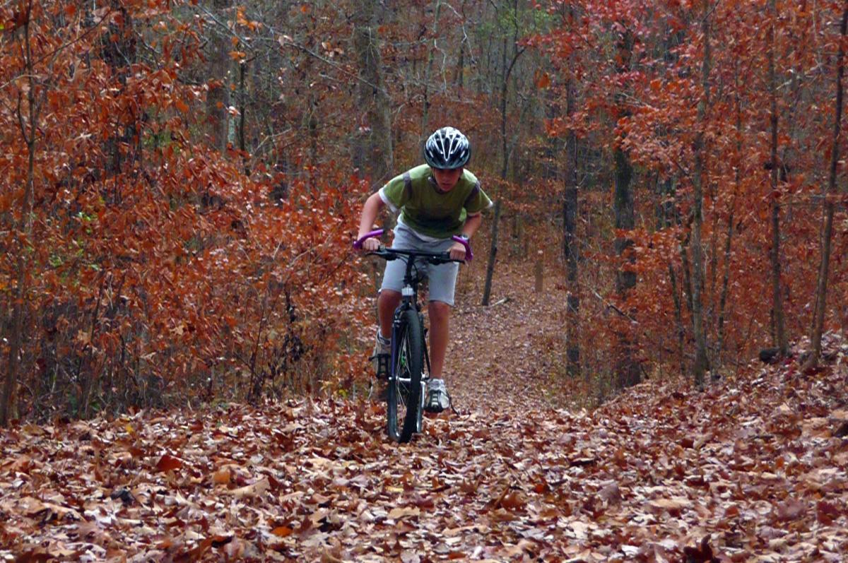 A young person riding a mountain bike on a forest trail covered with autumn leaves, surrounded by trees with orange and red foliage. The cyclist is leaning forward, focused on navigating the path. Yellow River mountain bike trail.