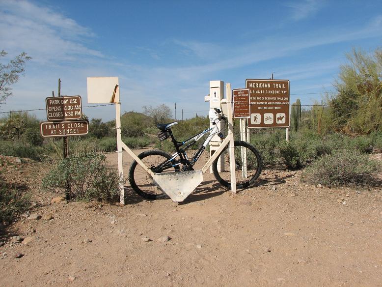 A mountain bike is parked at the entrance to the Meridian Trail, surrounded by desert vegetation. Two signs are visible: one indicating park hours of operation and the other detailing trail rules and information. The area is sunny with a clear blue sky in the background. Blevins Via Meridian mountain bike trail.