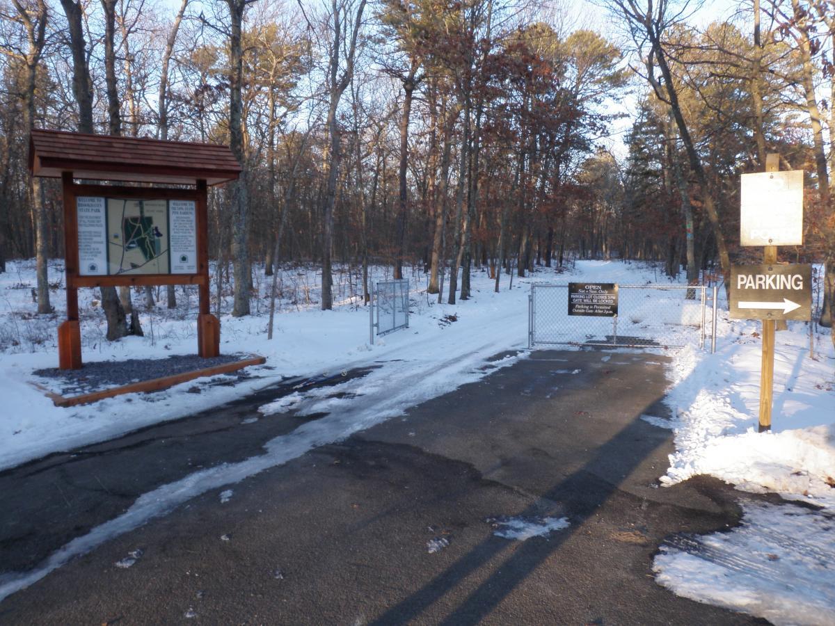 Park entrance with a trail map sign on the left and a parking sign directing to the right. Snow covers the ground, and trees are visible on either side of the paved roadway leading into the park. A gated entrance indicates open hours and park regulations. Brookhaven State Park mountain bike trail.