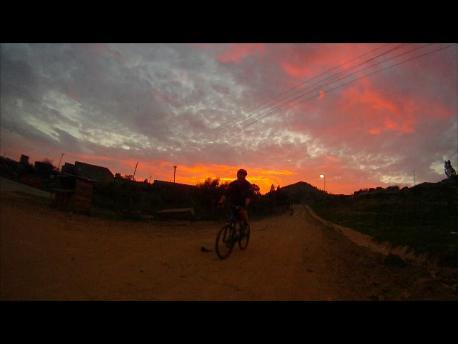 A cyclist riding along a dirt road at sunset, with vibrant orange and purple clouds in the sky and silhouettes of trees and houses in the background. Maseru Urban 12k mountain bike trail.