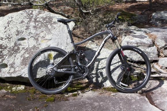 A mountain bike with a black and silver frame is leaning against a large rock in a natural outdoor setting. The bike features thick tires designed for rugged terrain and is surrounded by greenery and other rocks. Georgia International Horse Park mountain bike trail.