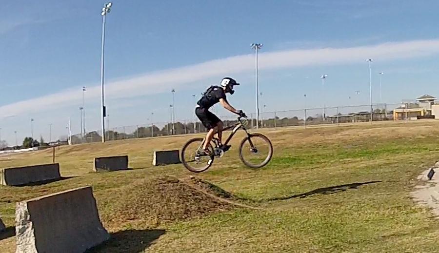 A person wearing a black helmet and clothing rides a mountain bike while performing a jump over a grassy slope. The background features a sports field with light poles and a clear blue sky. Loyce E. Harpe Park mountain bike trail.