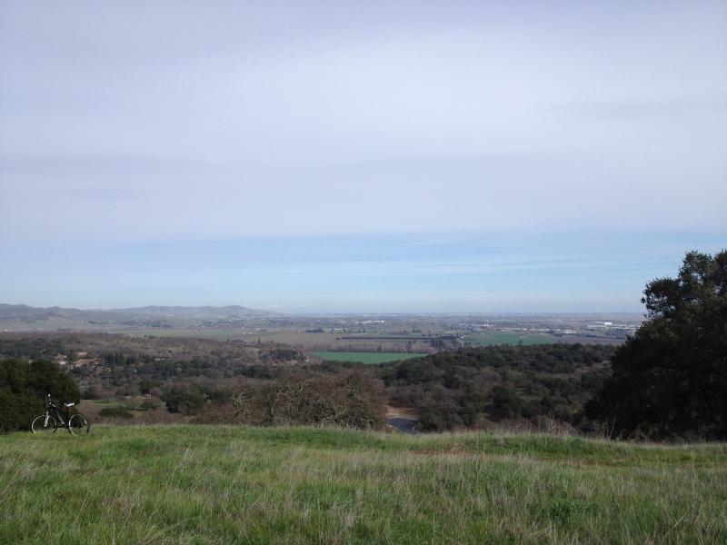 A scenic view from a hillside, showcasing a vast landscape of rolling hills and fields under a cloudy sky. A bicycle is parked on the grassy slope, adding a recreational element to the tranquil outdoor setting. Rockville Park mountain bike trail.