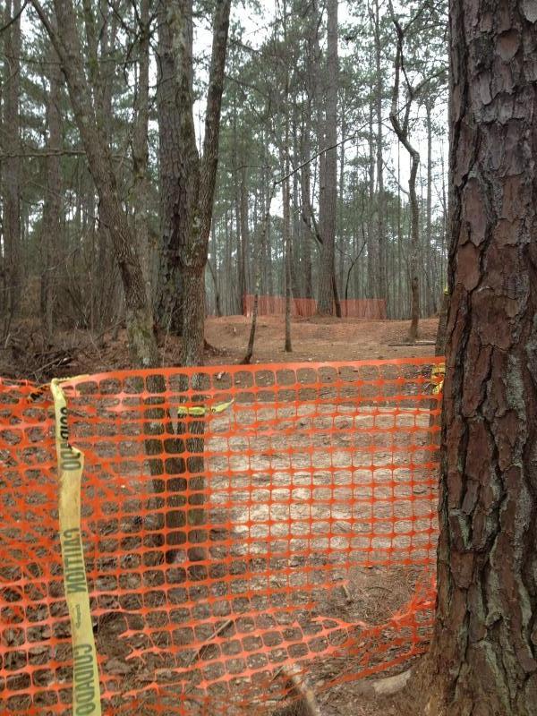 A forested area with tall trees, featuring a dirt path that is marked off with orange construction fencing and caution tape. The background shows more trees and a cleared section of the ground. The scene is overcast and indicates an area that may be under construction or restricted access. Quehl Holler mountain bike trail.