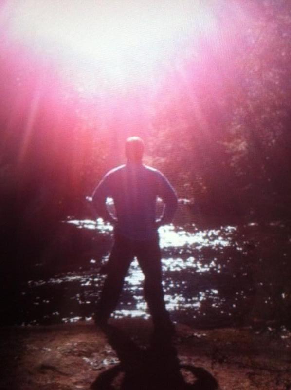 A silhouette of a person standing by a water's edge, facing a bright light source, possibly the sun, with shimmering reflections on the water surface. The surrounding environment appears to be natural, filled with trees and soft light filtering through. Cane Creek (sheltowee Trace Trail) mountain bike trail.