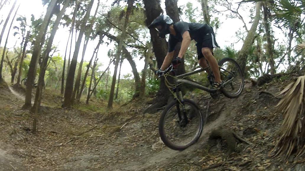 A mountain biker in a helmet and athletic gear jumping off a dirt ramp in a wooded area, surrounded by trees and foliage. The biker is mid-air, demonstrating a dynamic and adventurous pose while riding a mountain bike. Little Big Econ State Forest mountain bike trail.