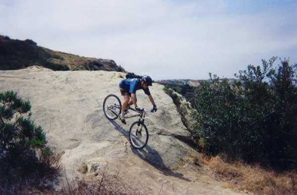 A mountain biker riding down a rocky hill on a sunny day, with hills and greenery in the background. Aliso and Wood Canyons Wilderness Park mountain bike trail.