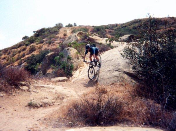 A mountain biker navigating a rocky trail on an uphill hillside. The scene features sparse vegetation and natural rock formations, with the biker in mid-action as he descends over a boulder surrounded by dry grass and shrubs. A clear sky is visible in the background. Aliso and Wood Canyons Wilderness Park mountain bike trail.