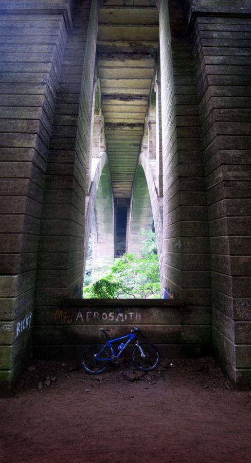 A blue bicycle resting on the ground beneath a large stone bridge. The bridge features arched supports, with greenery visible in the background. The walls of the bridge are covered in graffiti. The setting conveys a sense of tranquility and nature juxtaposed with urban structures. Wissahickon Valley Park mountain bike trail.
