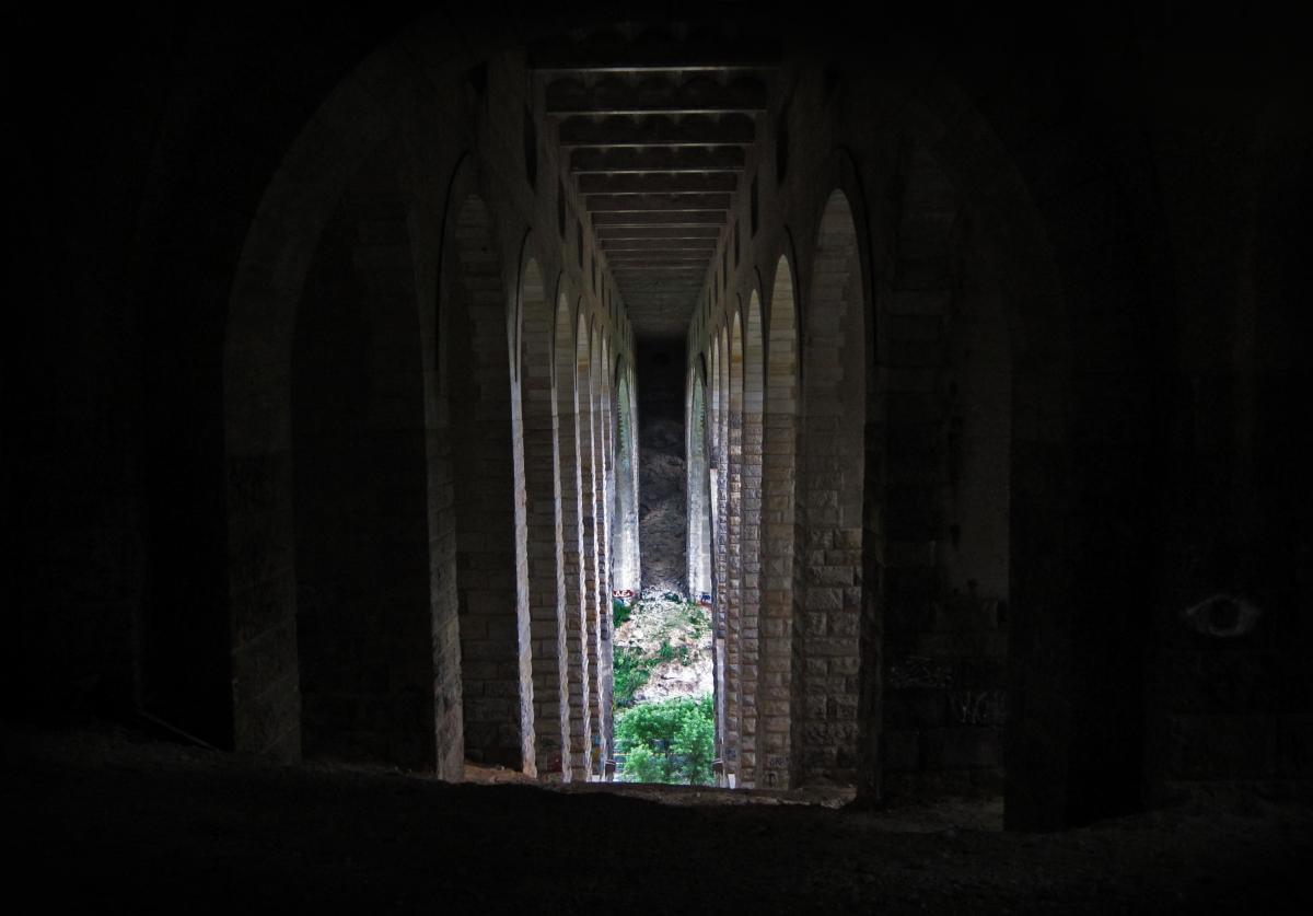 A dark corridor featuring arched stone supports, leading to a glimpse of green vegetation and a rocky backdrop beyond. The image highlights the contrast between light and shadow within the structure. Wissahickon Valley Park mountain bike trail.