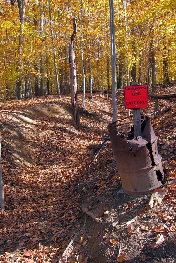 A scenic autumn landscape featuring a dirt trail surrounded by colorful fall foliage. A sign reading "Corkscrew Trail - EXIT ONLY" is visible next to a rusty, weathered barrel. Leaves cover the ground, indicating the season. The trail curves and leads off into the woods. Middle Run Natural Area mountain bike trail.