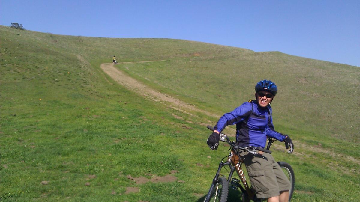 A person in a blue shirt and helmet is smiling while sitting on a mountain bike on a grassy trail. In the background, another cyclist is riding up a winding dirt path on a hillside under a clear blue sky. Havey Canyon Trail mountain bike trail.