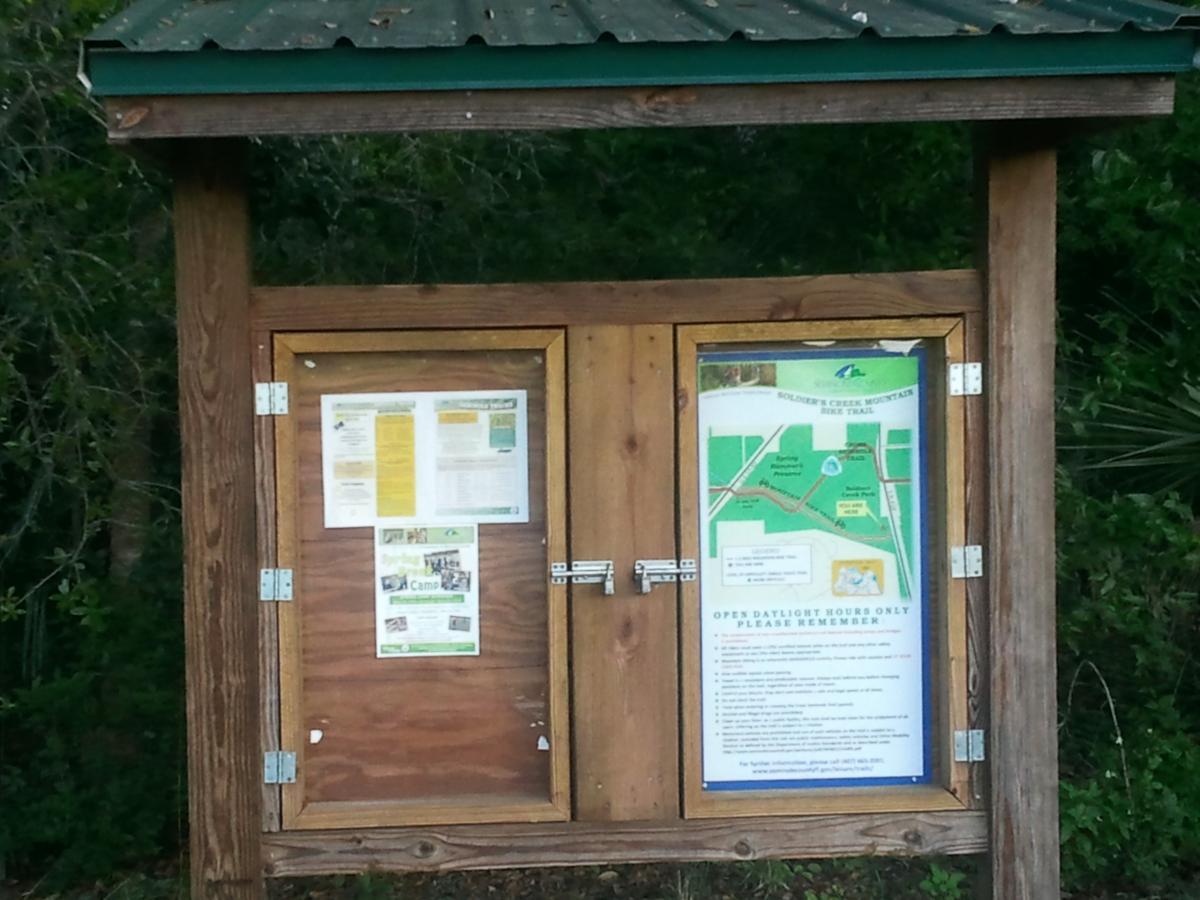 A wooden information kiosk with a green metal roof, featuring a display of maps and flyers on its front. The left side showcases various informational pamphlets, while the right side contains a map highlighting the Soldier's Creek Mountain Bike Trail, along with written guidelines and reminders for visitors. Surrounding vegetation is visible in the background. Soldier Creek Park mountain bike trail.