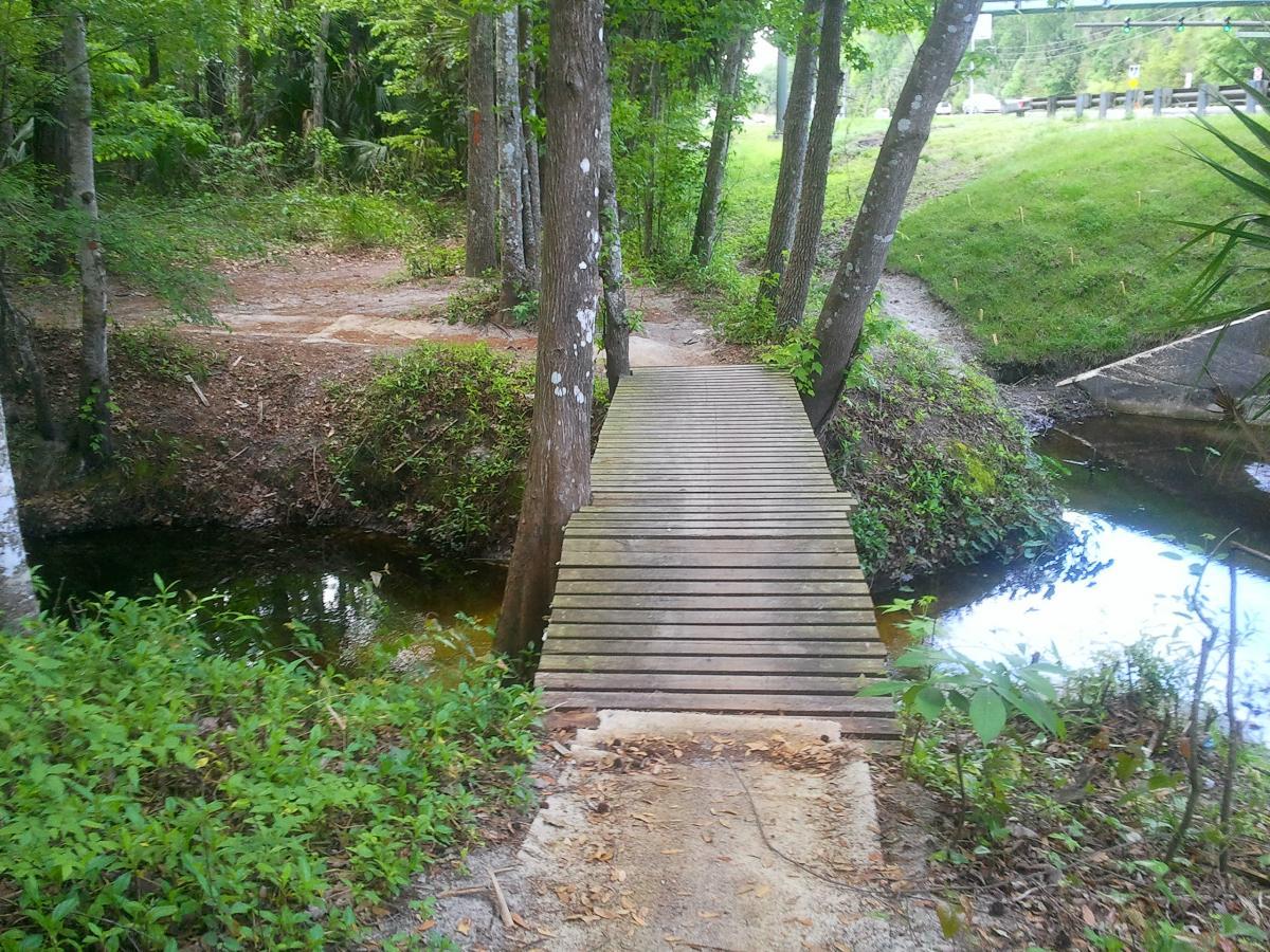 A wooden bridge spans a small stream surrounded by lush green foliage and trees. The area features a dirt path leading towards the bridge and is bordered by dense undergrowth. In the background, a grassy slope rises up with a road visible at the top, indicating the park's edge. Soldier Creek Park mountain bike trail.
