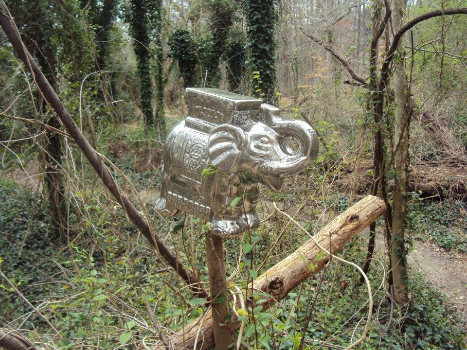 A shiny, metallic elephant sculpture perched on a fallen log in a wooded area, surrounded by trees and vines. Lake Maury mountain bike trail.