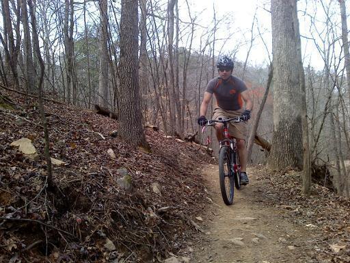 A mountain biker navigating a dirt trail through a wooded area, surrounded by trees and rocky terrain. The rider is wearing a helmet and a casual outfit, showcasing a focused expression as he rides along the winding path. Taylor Randahl Memorial Mountain Bike Trails At Olde Rope Mill Park mountain bike trail.