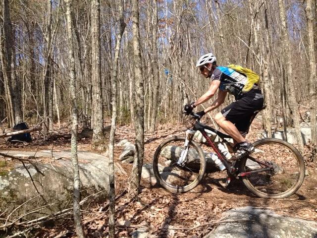 A mountain biker navigating a rocky trail in a dense forest, wearing a helmet and a backpack. The scene includes bare trees and fallen leaves on the ground, with the cyclist focused on maneuvering over the rocks. Five Points mountain bike trail.