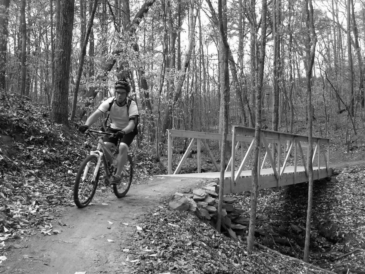A mountain biker rides along a dirt trail in a forested area, approaching a wooden bridge. The image is in black and white, capturing the rugged terrain and autumn foliage scattered on the ground. Trees line the path, creating a tranquil, outdoor scene. Taylor Randahl Memorial Mountain Bike Trails At Olde Rope Mill Park mountain bike trail.