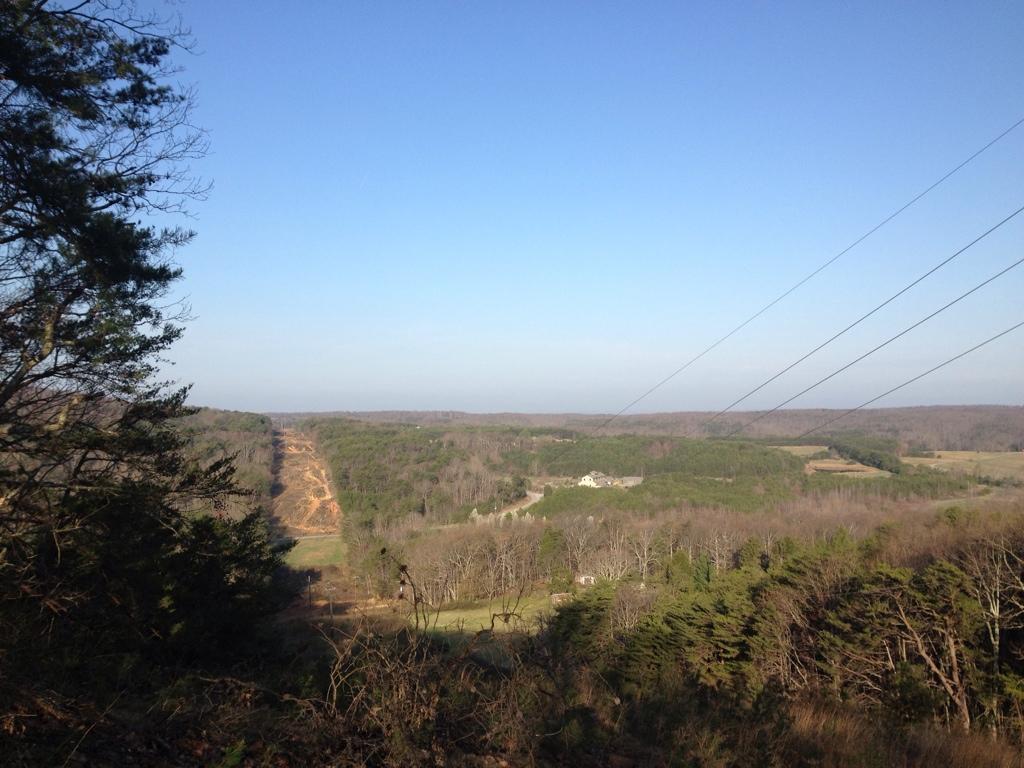 A panoramic view of a hilly landscape under a clear blue sky. The foreground features a mix of trees, while the background reveals rolling hills and a visible clearing, possibly indicating an area of land development. In the distance, a house is situated among the trees, and power lines stretch across the sky, highlighting the blend of natural and human elements in the scenery. Five Points mountain bike trail.