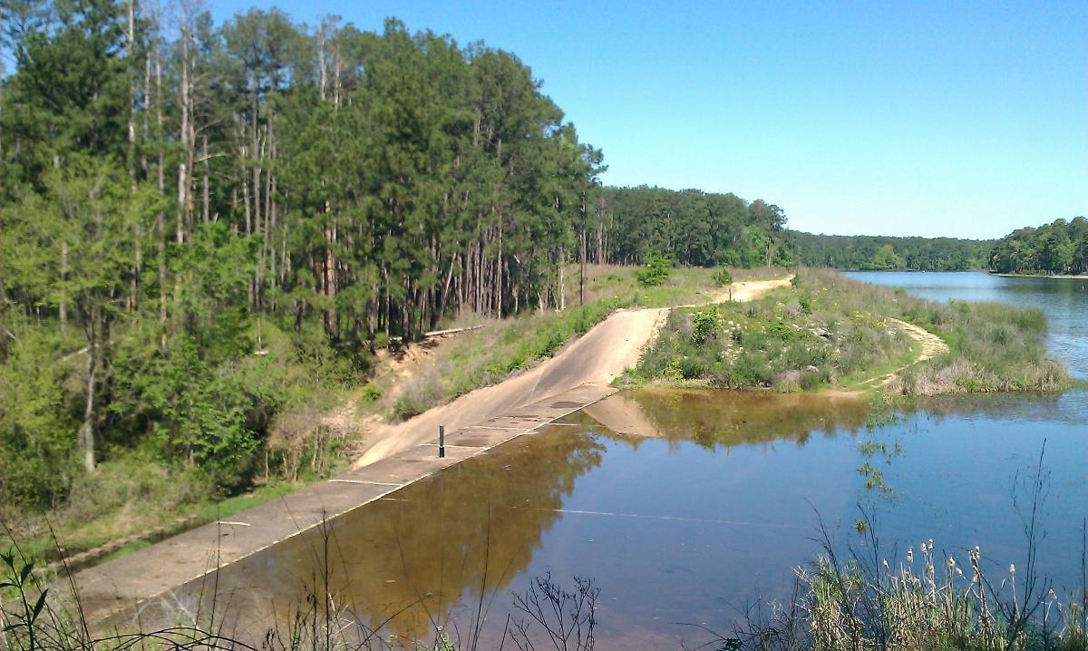 A serene landscape featuring a calm lake with a small, sloped boat ramp leading into the water. Surrounded by lush greenery and tall pine trees, the scene captures the tranquility of nature on a clear blue sky day. The reflection of the trees can be seen in the water, enhancing the peaceful atmosphere. Huntsville State Park mountain bike trail.