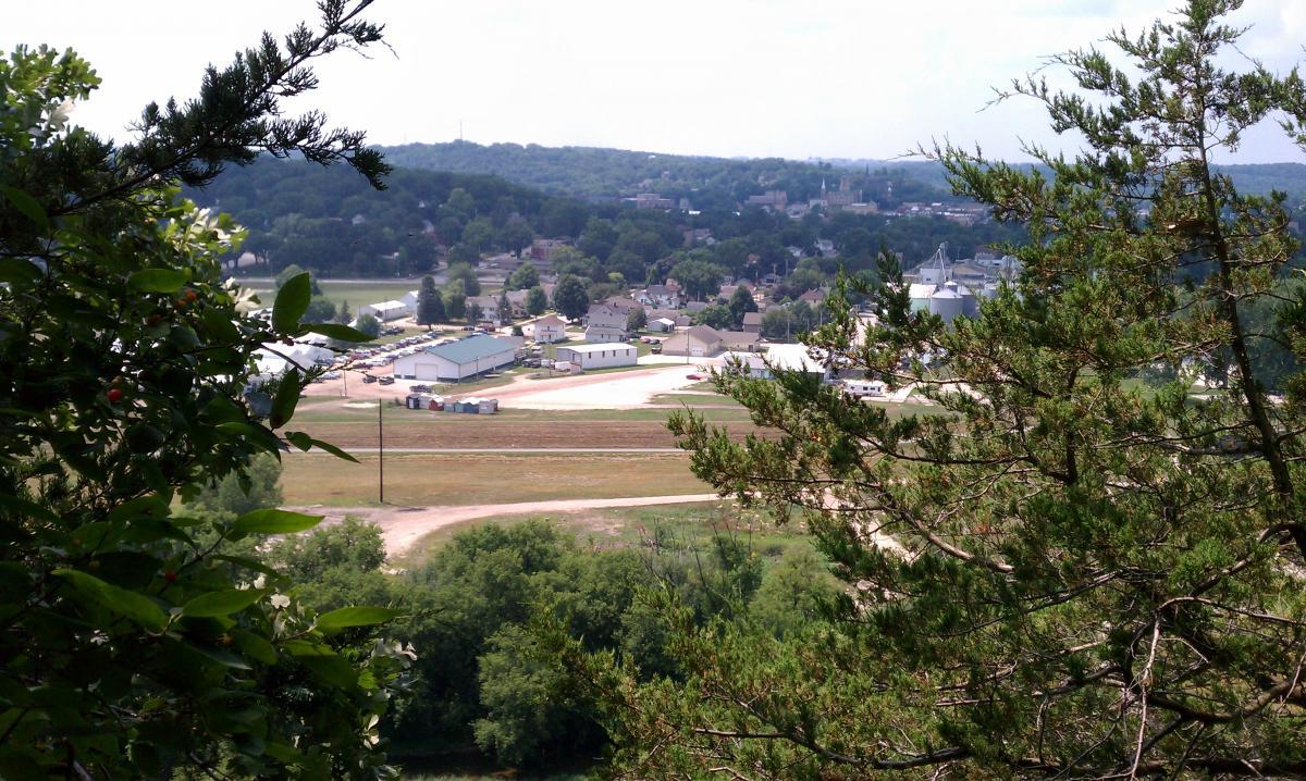 A panoramic view of a small town nestled in a lush green landscape, viewed from an elevated perspective. The scene includes various buildings, including houses and storage facilities, surrounded by trees and hills in the background under a partly cloudy sky. Decorah Mtb Trail System mountain bike trail.
