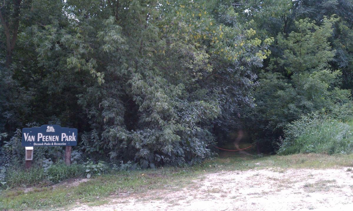 Sign for Van Peenen Park surrounded by dense greenery, with a dirt path leading into the woods. The sign includes the park's name and references Decorah Parks & Recreation. Decorah Mtb Trail System mountain bike trail.