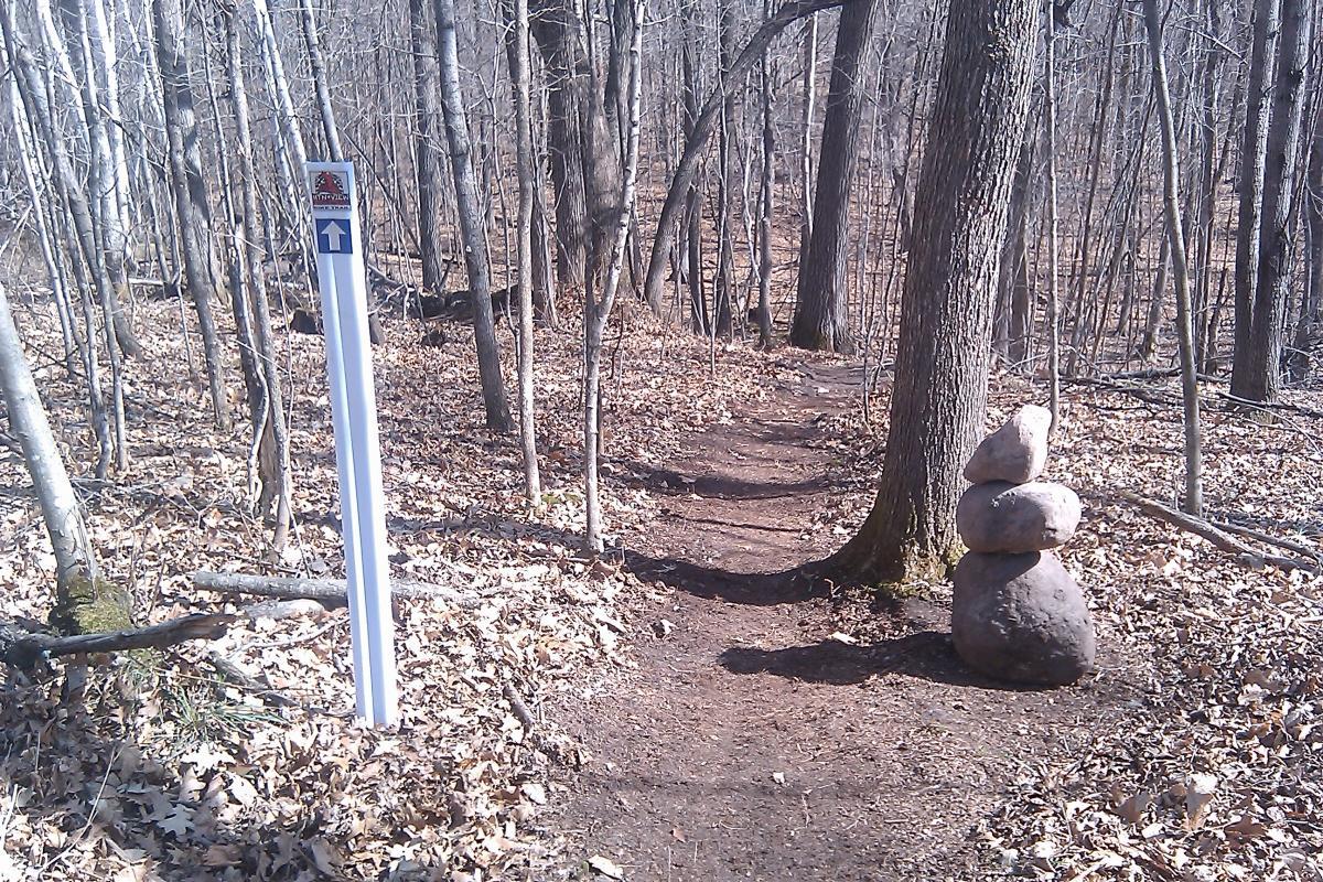 A hiking trail in a wooded area during early spring, featuring a directional sign with an arrow pointing upward and a stack of three smooth stones beside the path. The ground is covered with dry leaves, and the trees are bare, indicating a transition from winter to spring. Mountain View Singletrack mountain bike trail.