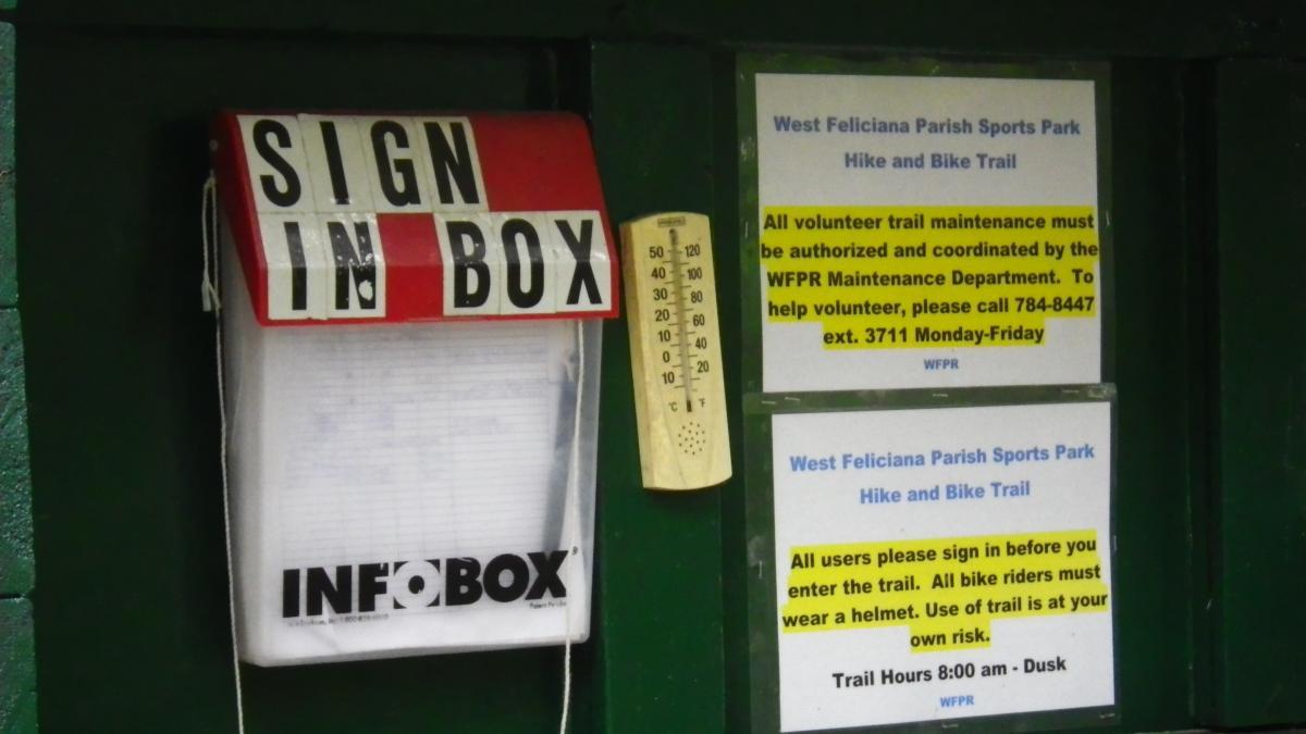 Sign-in box and information signs for the West Feliciana Parish Sports Park Hike and Bike Trail, displaying volunteer instructions and safety guidelines. A thermometer is also visible next to the sign-in box. The Beast mountain bike trail.