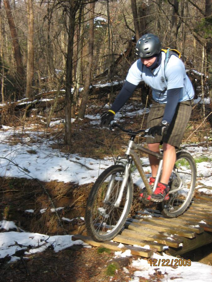 A cyclist riding a mountain bike over a wooden bridge in a snowy forest. The rider, wearing a helmet and a light blue shirt, appears focused as they navigate the trail. Surrounding trees and patches of snow create a wintery outdoor scene. Warriors' Path State Park mountain bike trail.