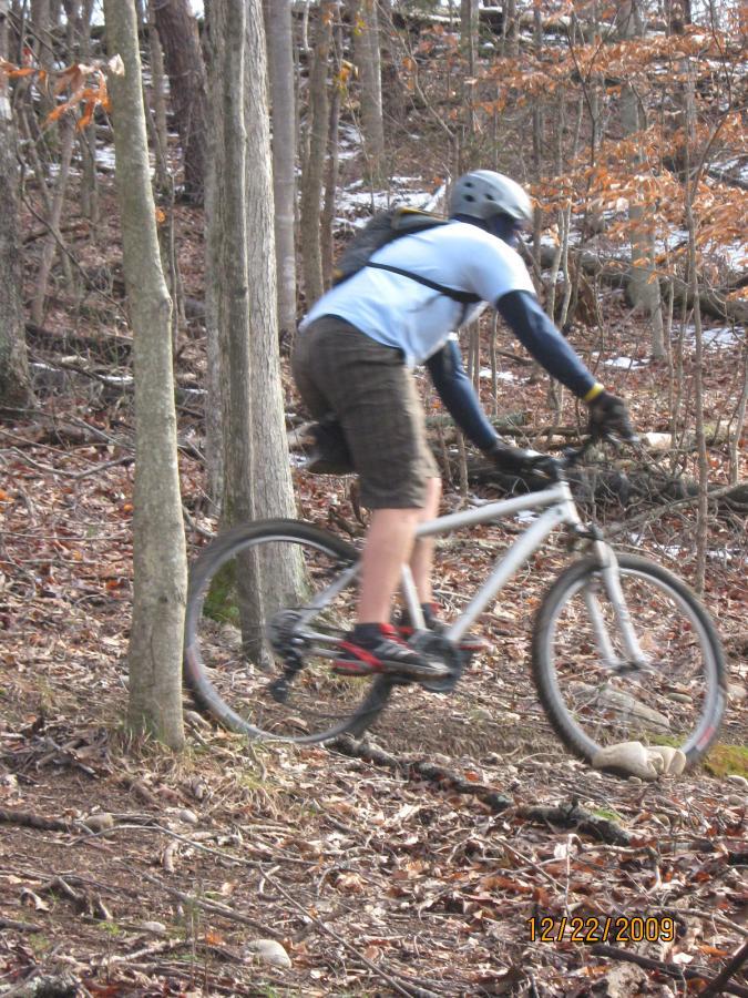 A mountain biker navigating a wooded trail, wearing a helmet and riding a white bike on a dirt path covered with leaves and small rocks. The setting features trees with bare branches and some fallen leaves, indicating a cool season. Warriors' Path State Park mountain bike trail.