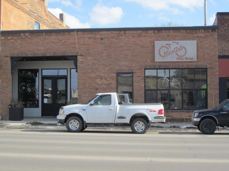 A brick storefront featuring the sign for "Nicollet Bike Shop." In front, a white pickup truck is parked next to a black truck. The building has large display windows showcasing bicycles, with a clear blue sky in the background. Snow is visible on the ground, indicating winter weather.