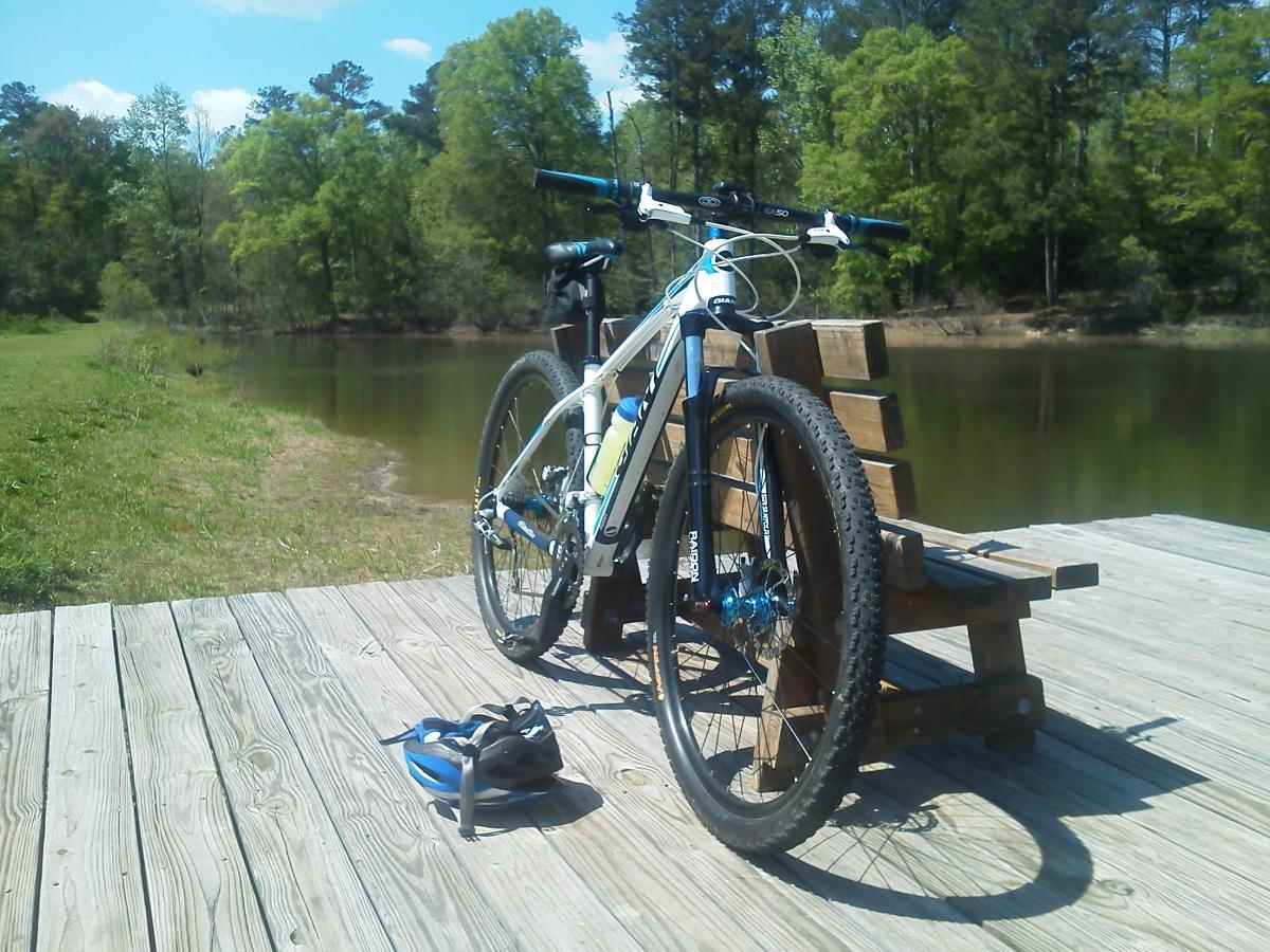 A mountain bike rests on a wooden dock by a calm pond, surrounded by greenery. A helmet sits on the dock nearby, and a bench is visible in the background. The sky is clear with a few clouds, indicating a sunny day. Children's Home / Pig Trail mountain bike trail.