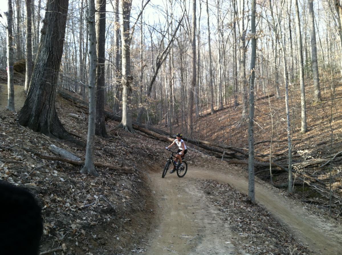 A person riding a mountain bike on a winding dirt trail in a wooded area. The landscape features tall, bare trees and scattered fallen branches, with dried leaves covering the ground. The scene is set in early spring, with no green foliage visible yet. Fountainhead Regional Park mountain bike trail.