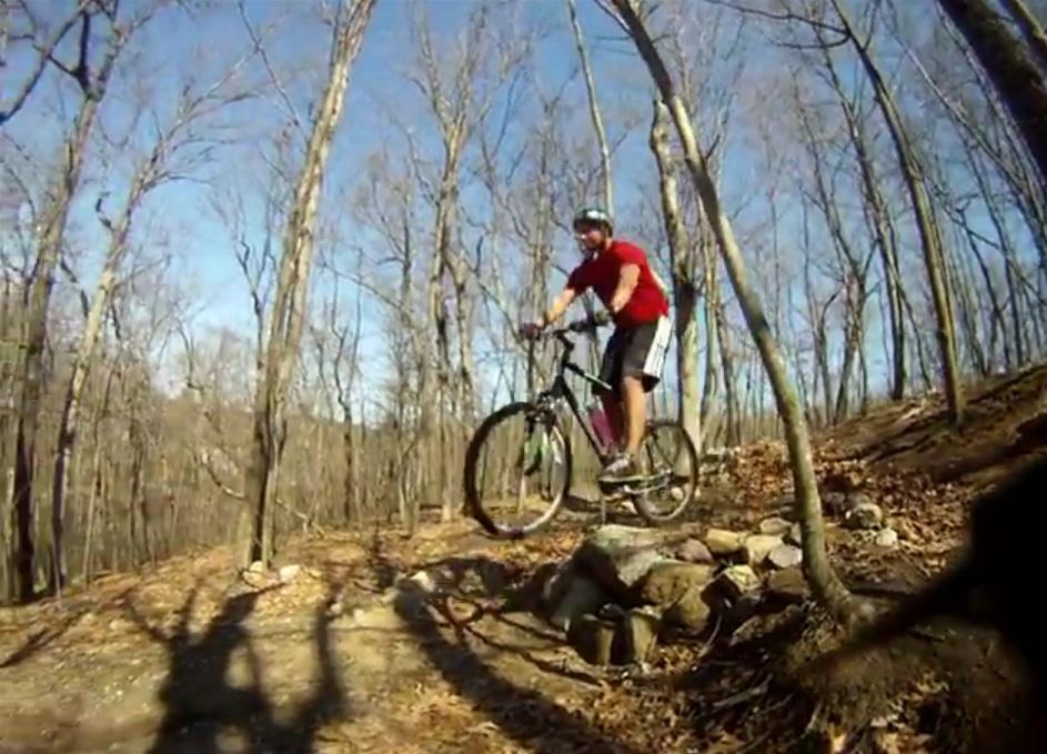 A person in a red shirt and black shorts riding a mountain bike, leaping off a small rocky outcrop in a wooded area with bare trees and a blue sky in the background. Yankee Springs mountain bike trail.