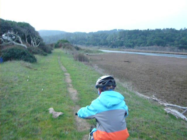 A child in a colorful jacket and helmet is riding a bicycle along a narrow dirt path beside a marshy area. Lush greenery and trees are visible in the background, with a river or waterway to the right. The scene suggests an outdoor adventure in a natural setting. Tomalas Bay Trail mountain bike trail.