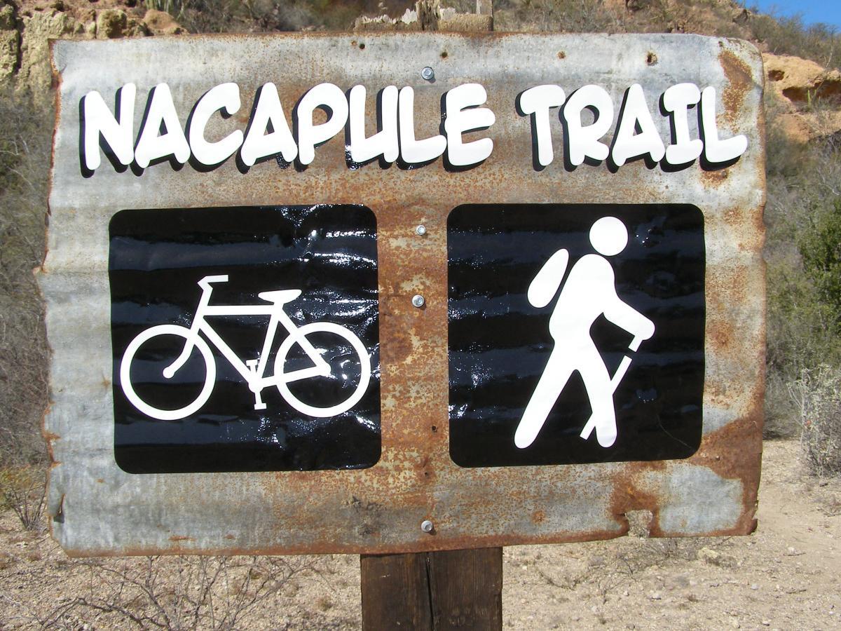 Sign for Nacapule Trail featuring icons for biking and hiking. The sign is weathered with a rusted metal background, showing symbols for a bicycle and a hiker on a dark background. Nacapule Trail mountain bike trail.