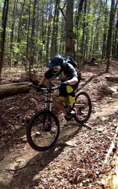 A person wearing a helmet and protective gear rides a mountain bike on a dirt trail through a forest. They are seen navigating around a fallen log, with trees and leaves surrounding the path. Big Creek mountain bike trail.