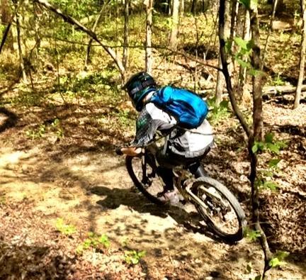 A person riding a mountain bike down a narrow trail in a wooded area. The cyclist is wearing a helmet and a blue backpack, navigating through the trees and foliage on a sunny day. Big Creek mountain bike trail.